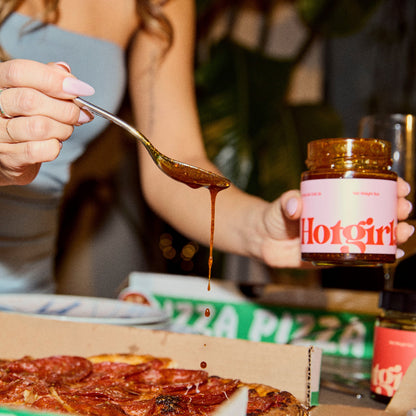 Person holding a spoon with hot sauce over a pizza, with 'Hotgirlsauce' chili oil in the background.