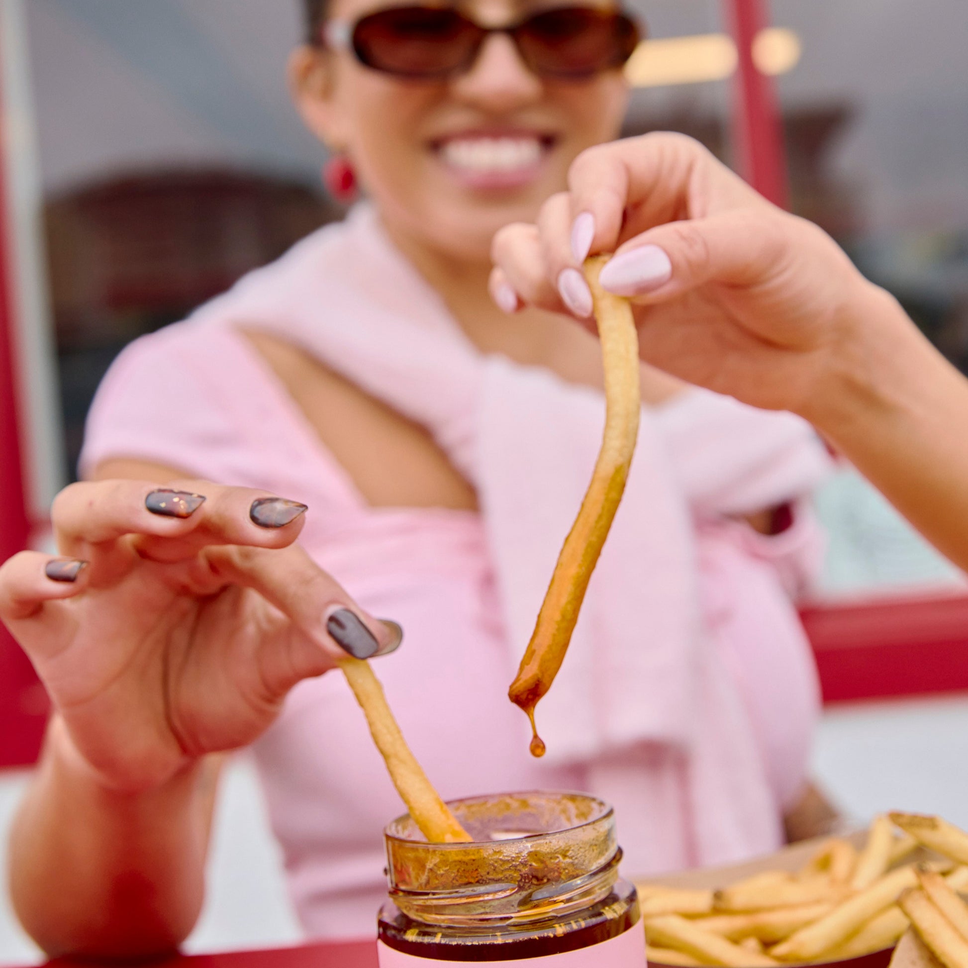Person dipping a fry into a jar of Hotgirlsauce chili oil hot sauce at a casual dining setting.

