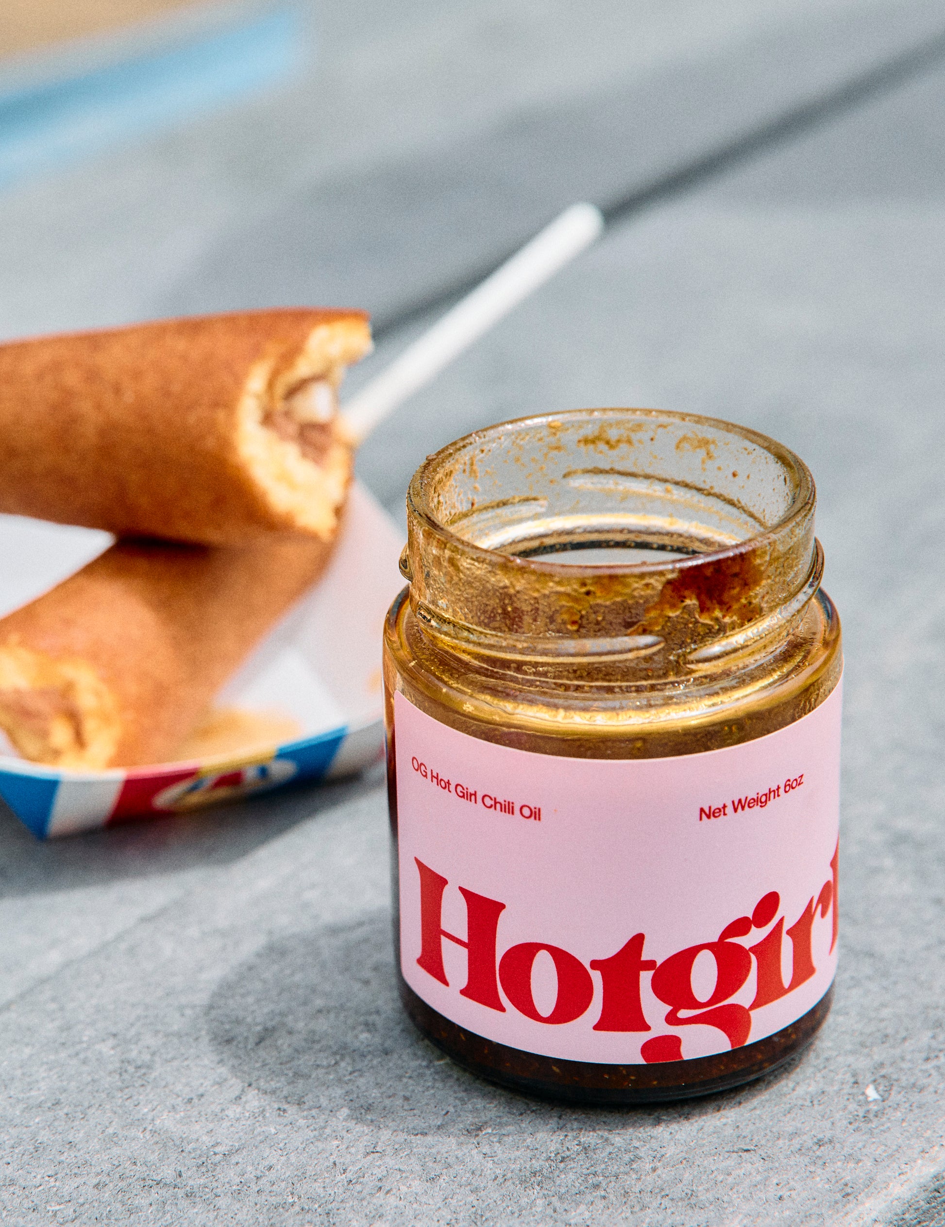 Jar of 'Hotgirlsauce' chili oil hot sauce on a gray surface with a corndog in the background

