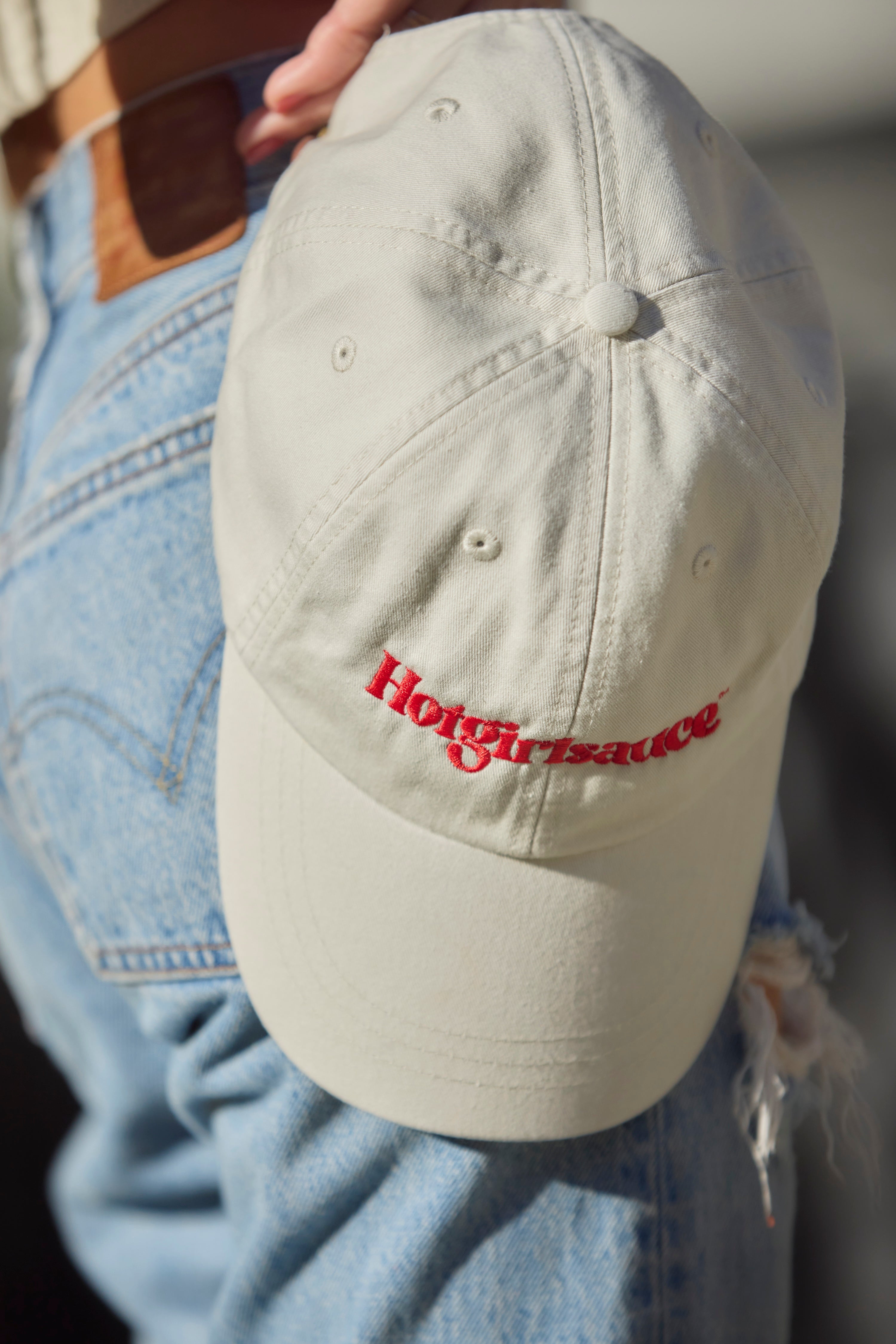 Close-up of Hotgirlsauce embroidered logo on cream dad hat in hands by hips